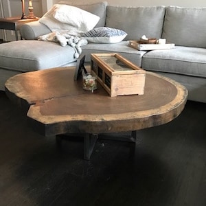 May include: A large, round wooden coffee table with a natural wood finish and a black metal base. The table is sitting on a dark hardwood floor.