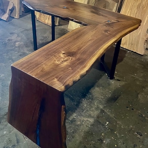 May include: A large, L-shaped desk made from a single piece of wood with a live edge. The desk has a dark brown wood finish and a blue epoxy resin inlay. The desk is supported by a black metal frame.
