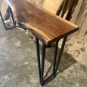 May include: A live edge console table with a rich brown wood top and black metal legs. The table's natural edge adds a unique, organic touch. The legs are made of black metal and are angled for stability.
