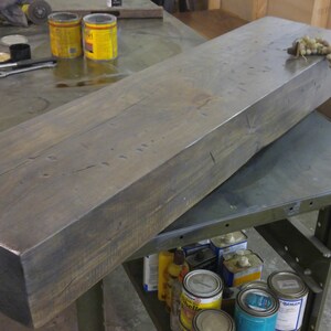 May include: A large, dark gray wooden beam with a rough, rustic texture. The beam is lying on a metal workbench with other items in the background.