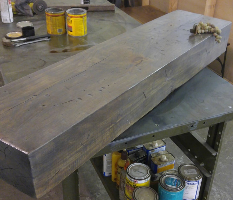 May include: A large, dark gray wooden beam with a rough, rustic texture. The beam is lying on a metal workbench with other items in the background.