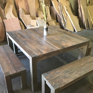 May include: A rustic wooden dining table with two matching benches. The table and benches are a dark gray color and have a distressed finish. The table is square and has a simple design. The benches are rectangular and have a slightly curved top. The table and benches are set against a background of stacked wood planks.