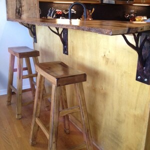 May include: Two wooden bar stools with a natural wood finish sit in front of a wooden countertop with a live edge. The countertop is supported by two black metal tree branch-shaped brackets.