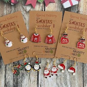 Peut inclure: Cinq paires de boucles d'oreilles à crochet dorées avec des breloques en émail sur le thème de Noël : bonhommes de neige, pulls à rennes, sacs cadeaux rouges avec texte Merry Christmas, pingouins et pères Noël sur des cartes brunes.