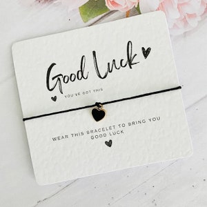 May include: A white card with black text that reads "Good Luck" and "You've got this". A black string bracelet with a gold heart charm is attached to the card. The text on the card also reads "Wear this bracelet to bring you good luck".