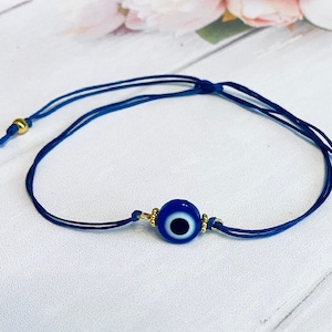 May include: A blue evil eye charm bracelet with a gold bead accent on a dark blue cord.