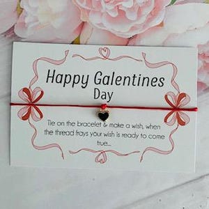 May include: A white card with the text "Happy Galentines Day" and a red string bracelet with a heart charm. The card has a decorative red border with ribbon and heart accents. The text below reads "Tie on the bracelet & make a wish, when the thread frays your wish is ready to come true..."