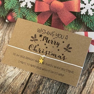 Puede incluir: Una tarjeta marrón con el texto "Wishing you a Merry Christmas" y una cuerda blanca con un dije de estrella dorada. El texto en la tarjeta dice "Tie on your bracelet and make a wish when you string starts to fray your wish is on its way!"