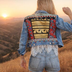 May include: A light-wash denim jacket with a colourful embroidered back panel featuring red floral patterns, gold accents, and intricate beadwork. The jacket is paired with denim shorts and a white lace top, set against a scenic sunset backdrop.