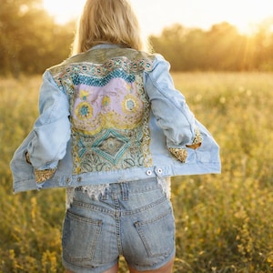 Könnte beinhalten: Hellblaue Jeansjacke mit bunt besticktem Rückenteil. Das Rückenteil hat ein Blumenmuster mit goldenen Pailletten-Akzenten an den Manschetten. Die Jacke wird mit Jeansshorts kombiniert. Die Person steht in einem Feld mit hohem Gras.