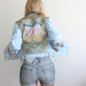 May include: A light blue denim jacket with a colourful patterned fabric patch on the back. The fabric patch features a paisley design with shades of pink, green, gold, and blue.