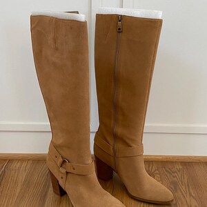 Puede incluir: Un par de botas de ante marr&oacute;n claro de altura de rodilla con un tac&oacute;n grueso y un detalle de hebilla en el eje.