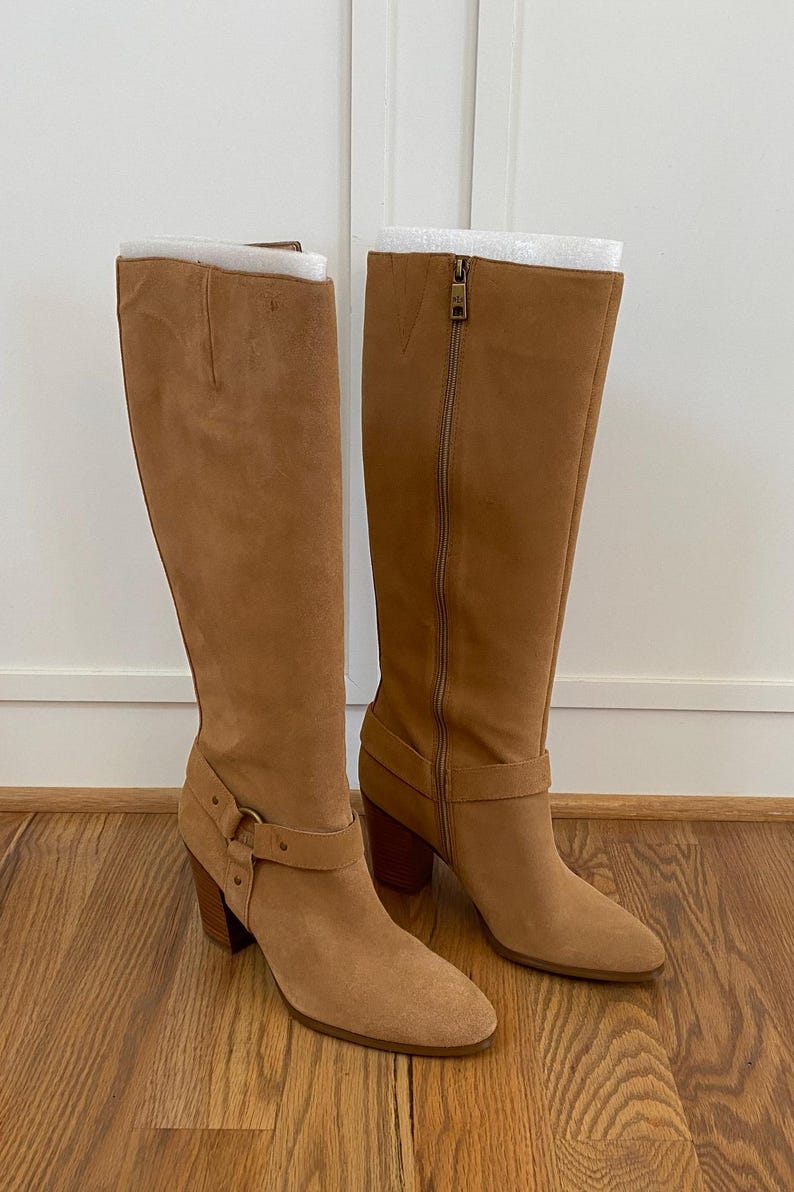 Puede incluir: Un par de botas de ante marr&oacute;n claro de altura de rodilla con un tac&oacute;n grueso y un detalle de hebilla en el eje.
