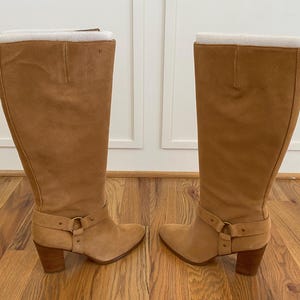 Puede incluir: Un par de botas de ante marr&oacute;n de ca&ntilde;a alta con tac&oacute;n grueso y detalle de hebilla en el eje.