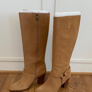 Puede incluir: Un par de botas de ante beige de ca&ntilde;a alta con un detalle de hebilla en una bota y una cremallera en la otra. Las botas tienen un tac&oacute;n de bloque.