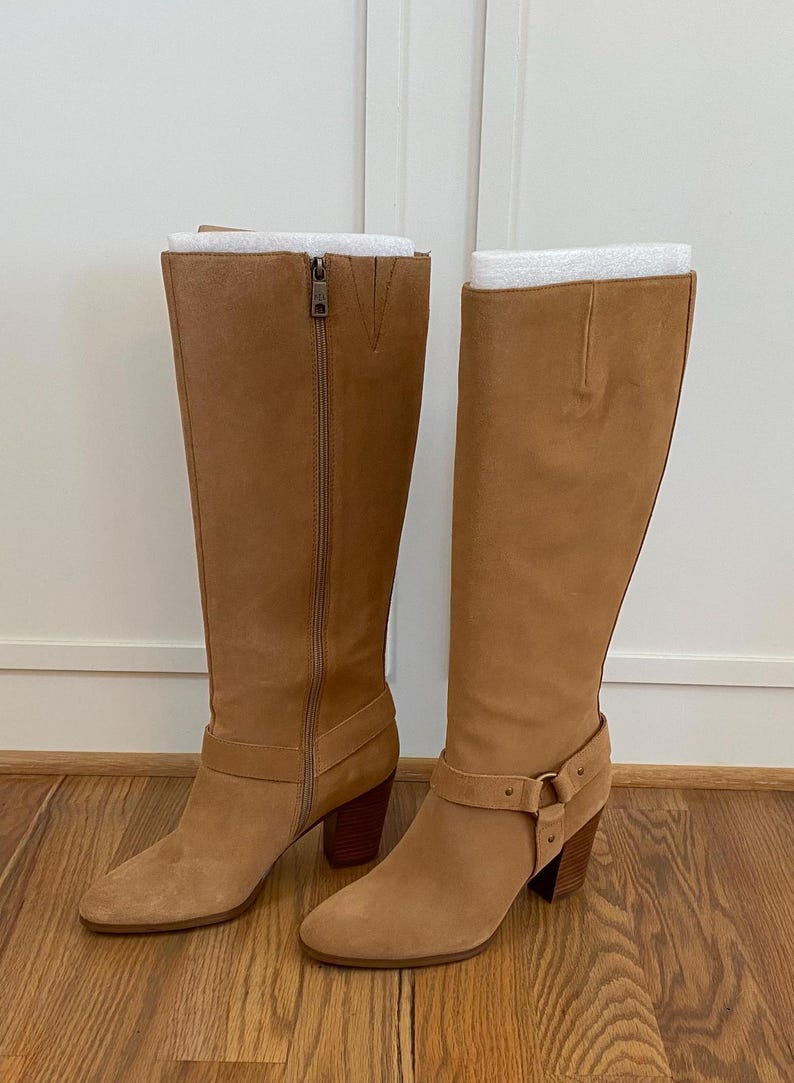 Puede incluir: Un par de botas de ante beige de ca&ntilde;a alta con un detalle de hebilla en una bota y una cremallera en la otra. Las botas tienen un tac&oacute;n de bloque.