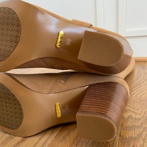 Puede incluir: Un par de botas de ante marr&oacute;n hasta la rodilla con tac&oacute;n de bloque. Las botas tienen una etiqueta dorada en el lateral con la talla "9.5B" impresa en ella.