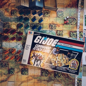 May include: A vintage G.I. Joe Commando Attack Game board game with a colorful box featuring a star and the text "G.I. Joe Commando Attack Game - A Real American Hero". The game board is partially visible with a green river, brown buildings, and red and blue game pieces.