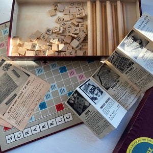 May include: A vintage Scrabble game set with a wooden game board, letter tiles, and instruction booklets. The game board is red and has a grid pattern. The letter tiles are made of wood and have black letters on them. The instruction booklets are folded and have black text on a white background.