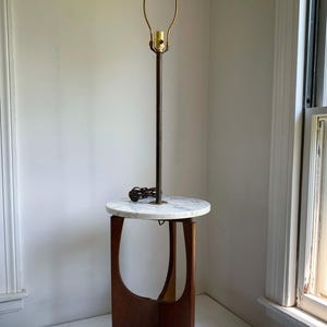 May include: A floor lamp with a circular white marble tabletop integrated into a brown wooden base. The lamp features a tall, thin, dark metal stem with a brass socket and a harp at the top. A brown electrical cord is coiled on the marble surface.