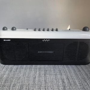 May include: A vintage Sharp boombox, featuring a black and white design. The radio has a cassette player and multiple control knobs. The word "SHARP" is visible on the front. The boombox sits on a gray surface.