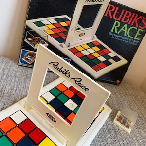 May include: A Rubik's Race game with a white frame and colorful squares. The game board is open, displaying a grid of colored squares. The box is visible in the background, with the text "Rubik's Race" and "The fastest game in town!"