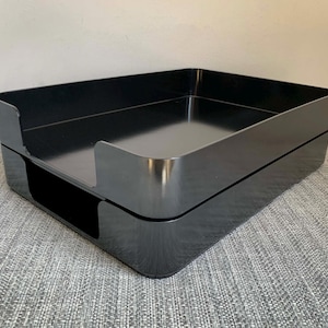 May include: A glossy black desk organizer with a rectangular shape. The organizer has a two-tiered design with rounded corners and a smooth, reflective surface. The top tier has a cutout for easy access. Ideal for office or home organization.