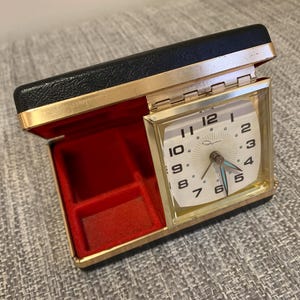 May include: A vintage travel alarm clock in a black and gold case. The clock face is white with black numerals and hands. The interior is lined with red velvet. The case is open, revealing the clock face.