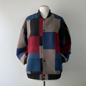 May include: A patchwork bomber jacket in a variety of colors, including red, blue, black, and tan. The jacket has a black collar, cuffs, and hem. It is buttoned up and displayed on a mannequin.