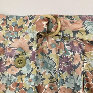 Puede incluir: Primer plano de tela con estampado floral con una trabilla beige y una hebilla grande y redonda de color beige. La tela presenta un estampado de flores en tonos rosa, morado, verde y marr&oacute;n. Se ve un bot&oacute;n en el lado izquierdo.