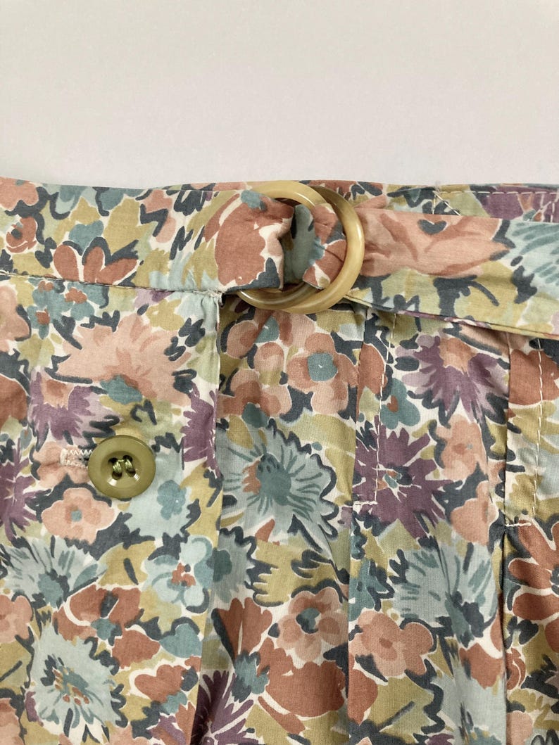 Puede incluir: Primer plano de tela con estampado floral con una trabilla beige y una hebilla grande y redonda de color beige. La tela presenta un estampado de flores en tonos rosa, morado, verde y marr&oacute;n. Se ve un bot&oacute;n en el lado izquierdo.