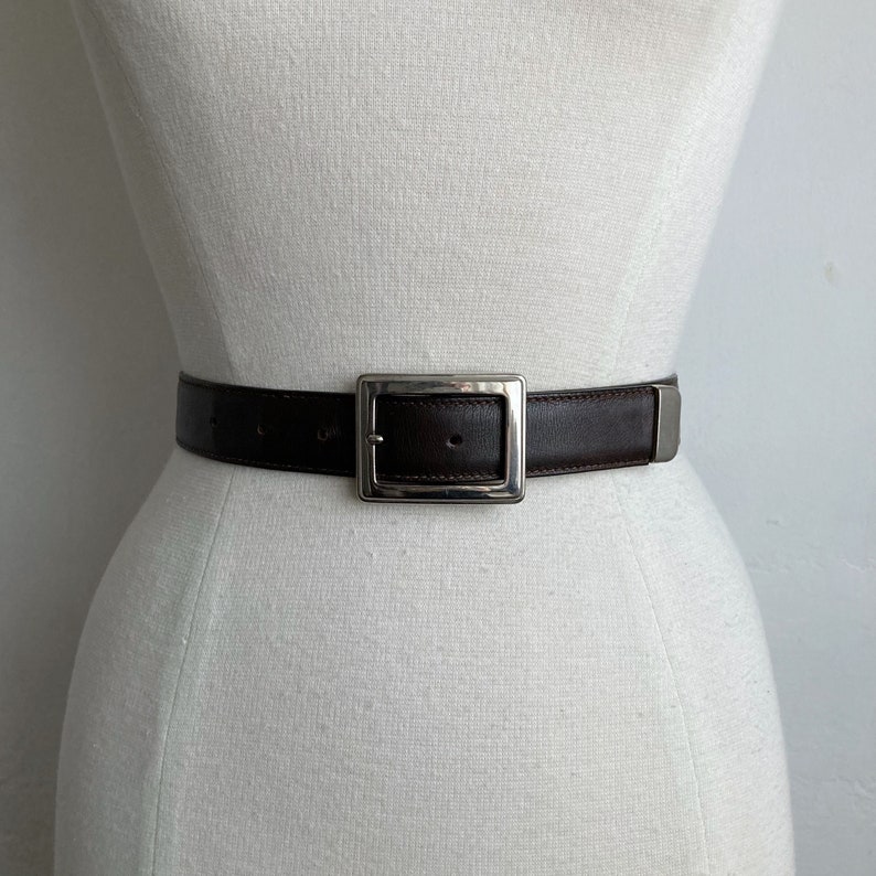 May include: A dark brown belt with a large, square, silver-coloured buckle. The belt is displayed on a white mannequin. The buckle is a prominent feature, contrasting with the dark leather. This belt is a classic accessory.