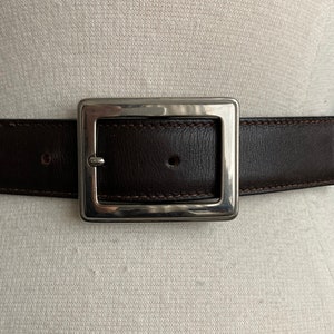 May include: A dark brown leather belt with a large, square, silver-toned buckle. The belt is stitched with a lighter brown thread and has a matching silver-toned end piece. The belt is displayed on a mannequin.