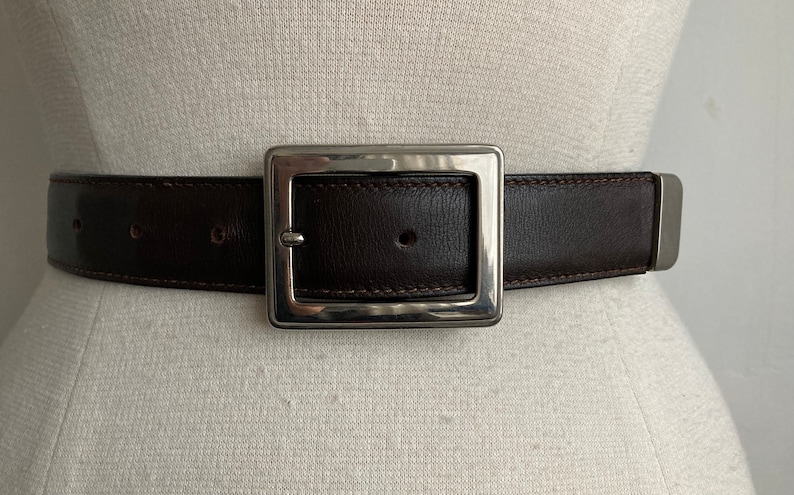 May include: A dark brown leather belt with a large, square, silver-toned buckle. The belt is stitched with a lighter brown thread and has a matching silver-toned end piece. The belt is displayed on a mannequin.