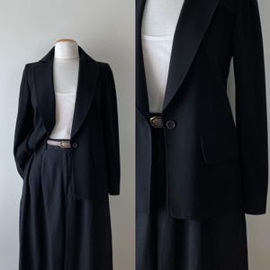 May include: A black blazer and matching pants are displayed on a mannequin. The blazer has a notched lapel, a single button closure, and two front pockets. A white tank top and a thin belt are visible. The pants are high-waisted and wide-legged.