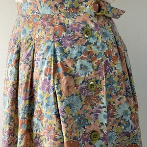 Puede incluir: Falda de cintura alta con estampado floral en tonos azules, melocot&oacute;n, morado y amarillo. La falda tiene cierre de botones y cintur&oacute;n con hebilla circular. La tela parece ser de algod&oacute;n o un material ligero similar.
