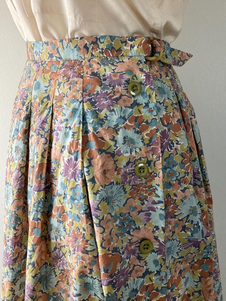 Puede incluir: Falda de cintura alta con estampado floral en tonos azules, melocot&oacute;n, morado y amarillo. La falda tiene cierre de botones y cintur&oacute;n con hebilla circular. La tela parece ser de algod&oacute;n o un material ligero similar.