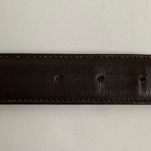 May include: A dark brown leather belt with a silver-coloured rectangular buckle. The belt has several evenly spaced holes and is stitched with a lighter brown thread. The belt is approximately 3.2 cm wide.