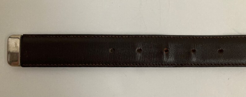 May include: A dark brown leather belt with a silver-coloured rectangular buckle. The belt has several evenly spaced holes and is stitched with a lighter brown thread. The belt is approximately 3.2 cm wide.