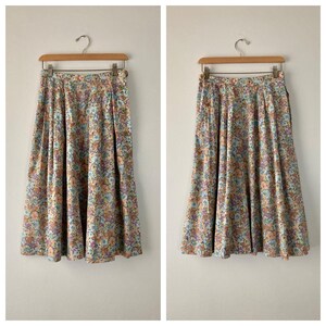 Puede incluir: Dos faldas con estampado floral colgadas de perchas de madera. Las faldas tienen un fondo azul claro con un estampado de peque&ntilde;as flores en tonos melocot&oacute;n, morado y verde. Las faldas son plisadas y de largo midi.