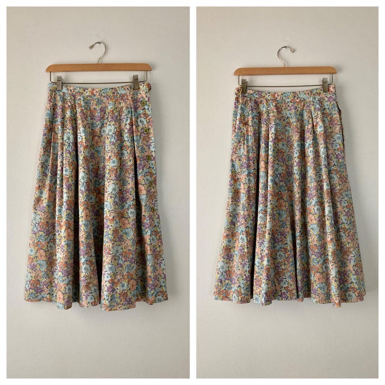 Puede incluir: Dos faldas con estampado floral colgadas de perchas de madera. Las faldas tienen un fondo azul claro con un estampado de peque&ntilde;as flores en tonos melocot&oacute;n, morado y verde. Las faldas son plisadas y de largo midi.