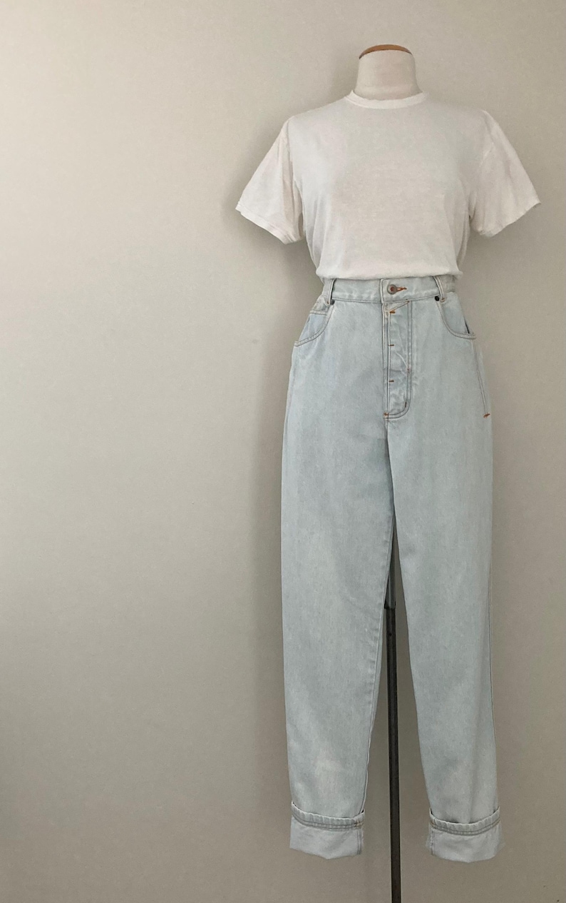 May include: A white t-shirt and light blue high-waisted jeans displayed on a mannequin. The jeans have a button fly and cuffed hems. The outfit is presented against a neutral background.