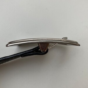 May include: A silver belt buckle with a curved rectangular shape. It is attached to a black leather belt. The buckle has a simple, elegant design. The belt is black.