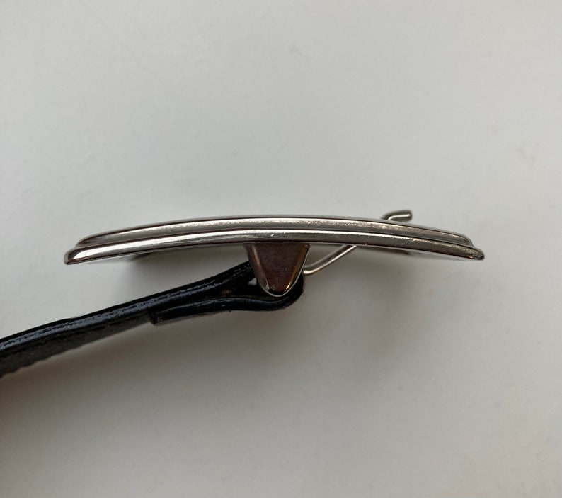 May include: A silver belt buckle with a curved rectangular shape. It is attached to a black leather belt. The buckle has a simple, elegant design. The belt is black.