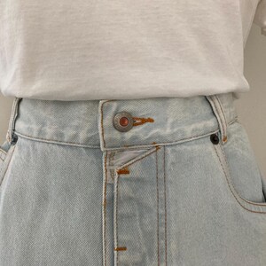 May include: Close-up of a light blue denim skirt with a button closure and pockets. The skirt is paired with a white t-shirt. The denim skirt has visible stitching and a classic design.