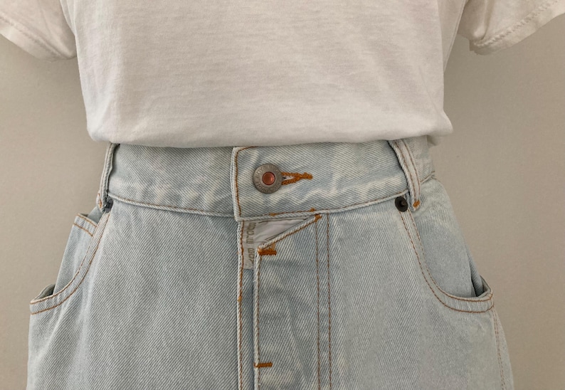 May include: Close-up of a light blue denim skirt with a button closure and pockets. The skirt is paired with a white t-shirt. The denim skirt has visible stitching and a classic design.
