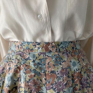 Puede incluir: Una camisa color crema con botones combinada con una falda con estampado floral. La falda presenta un estampado de flores en tonos azules, marrones y morados. El conjunto es de estilo cl&aacute;sico y femenino.
