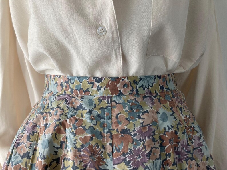 Puede incluir: Una camisa color crema con botones combinada con una falda con estampado floral. La falda presenta un estampado de flores en tonos azules, marrones y morados. El conjunto es de estilo cl&aacute;sico y femenino.