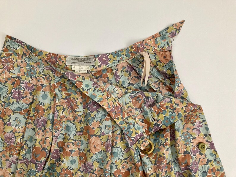 Puede incluir: Primer plano de una falda con estampado floral con dise&ntilde;o envolvente. La falda presenta una mezcla de colores, incluyendo flores melocot&oacute;n, azules y moradas. Tiene cierre de bot&oacute;n y detalle de anillo de madera. La tela parece ligera.