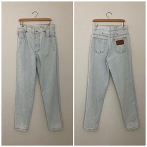 May include: Light blue denim jeans with a high waist and straight leg. The jeans have a button closure, pockets, and a brown leather patch on the back. The jeans are hanging on a wooden hanger.